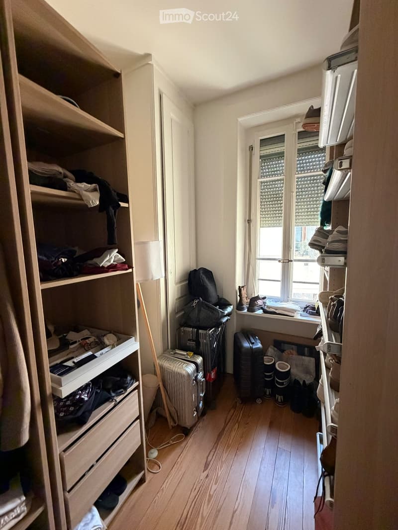 A narrow hallway with wooden flooring, built-in storage units on both sides, and a window at the end. Various items such as luggage, shoes, and clothing are placed along the walls.