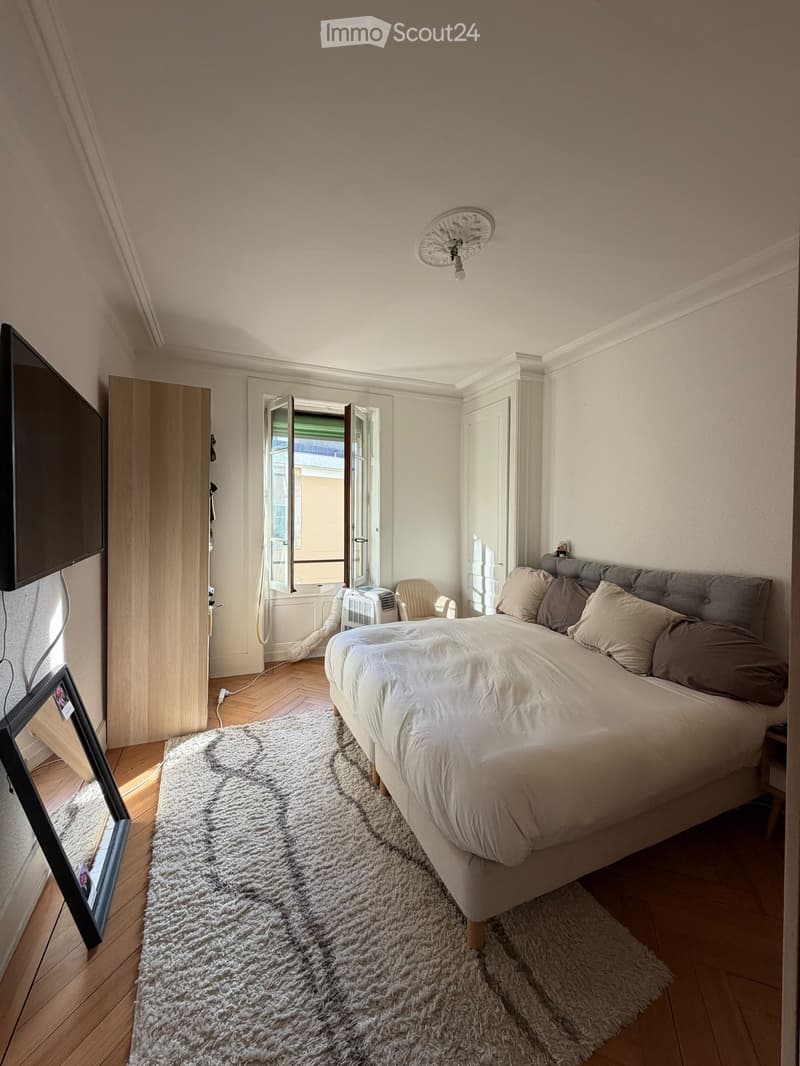 Cozy bedroom with a large bed, pillows, a fluffy rug, a chair, a screen, a wardrobe, and a fan. The room has wooden flooring and large windows with open shutters.