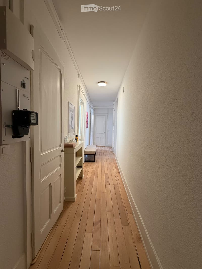 A narrow hallway with wooden flooring, white walls, and a small bench near a door at the end. A small table with items sits against one wall, and a light fixture is mounted on the ceiling.