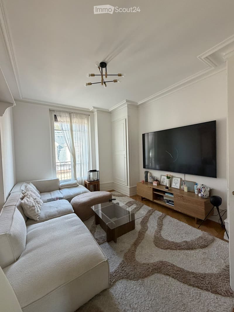 Modern living room with a grey sectional sofa, glass coffee table, ottoman, wooden side table, TV, and decorative items on a shelf.