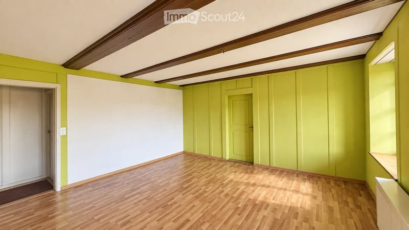Empty room with wooden flooring, exposed wooden beams on the ceiling, and green and white walls. There are two doors and a small shelf on the right side.