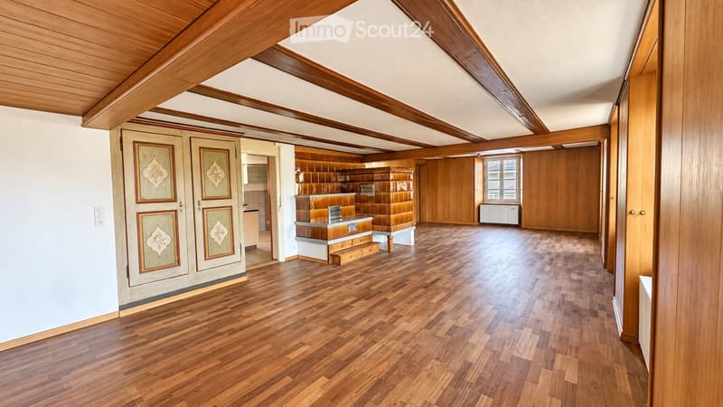 Spacious, unfurnished room with wooden flooring, exposed wooden beams on the ceiling, and a small kitchen area visible in the background.