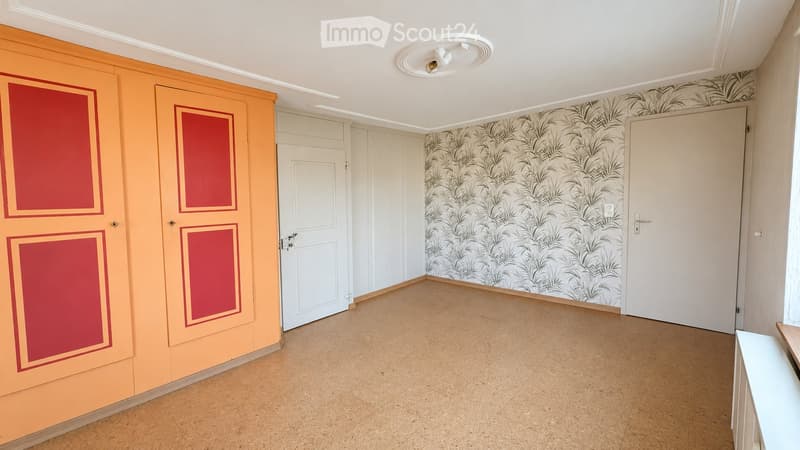 Empty room with light-colored walls, patterned wallpaper on one wall, and a wooden floor. Features include a white ceiling light fixture and several doors.