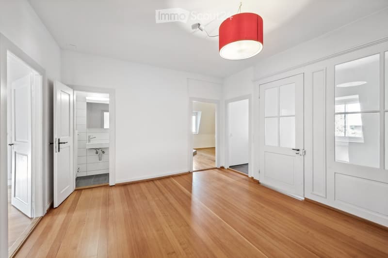Bright hallway with wooden flooring, white walls, and several doors leading to different rooms, including a bathroom with a sink and mirror visible through an open door.