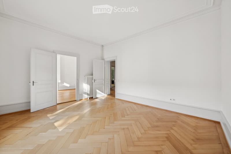Empty room with white walls, wooden herringbone floor, and open doors leading to other rooms.
