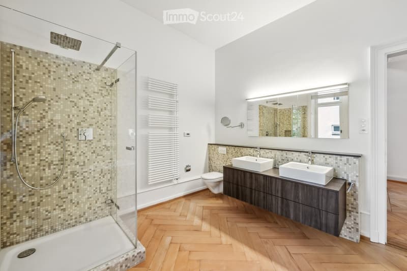 Modern bathroom with a glass-enclosed shower, double sink vanity, toilet, and a large mirror above the sinks. The floor is wooden, and the walls feature a mix of tiles and white paint.