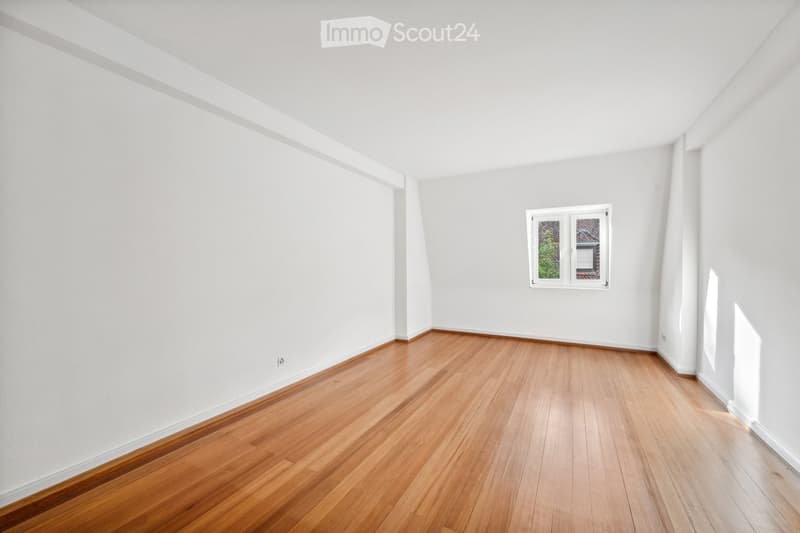 Empty room with wooden flooring and white walls, featuring a single window with a view of greenery outside.
