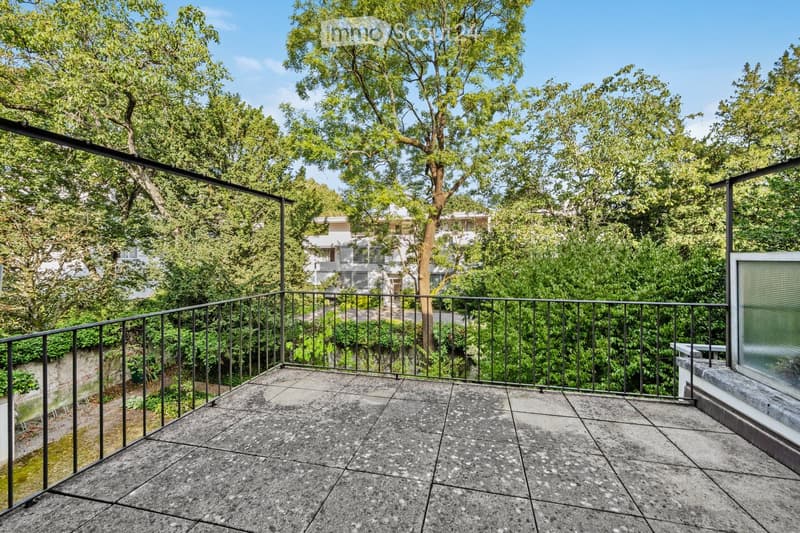 Balcony with a modern black metal railing, tiled concrete floor, and lush green trees in the background.