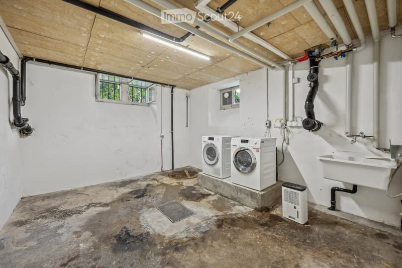 Utility room with white washing machine, dryer, sink, and air conditioner. Exposed pipes and concrete floor.