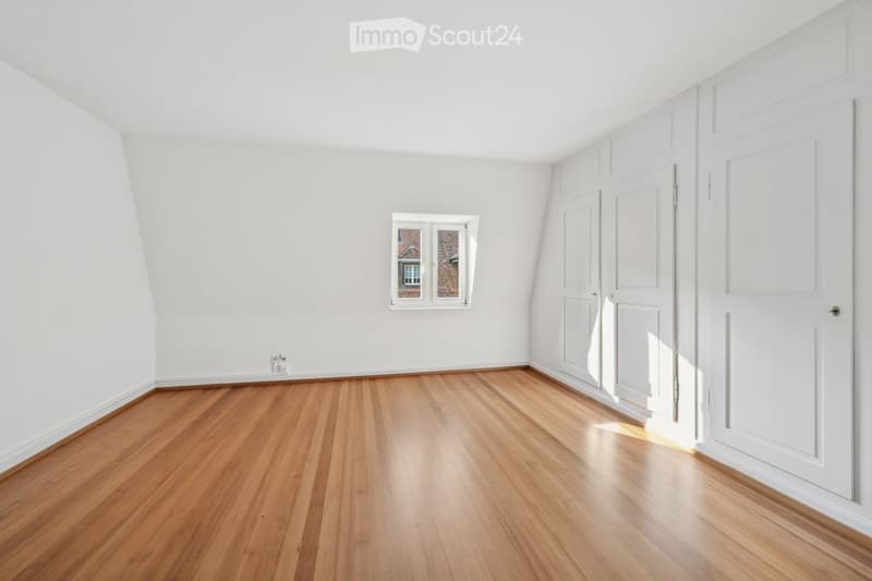 Empty room with wooden flooring, white walls, and a small window showing a view of a neighboring building.