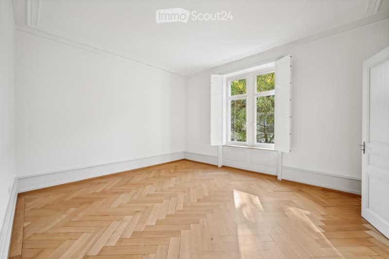 Empty room with white walls, wooden herringbone flooring, and large windows with shutters open to natural light.