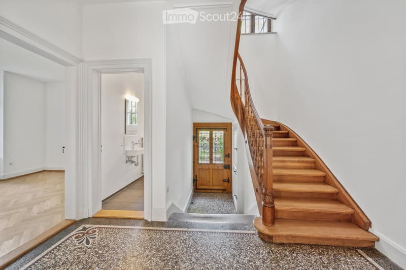 Elegant hallway with wooden staircase, polished wooden floors, white walls, and a small sink area visible in the background.