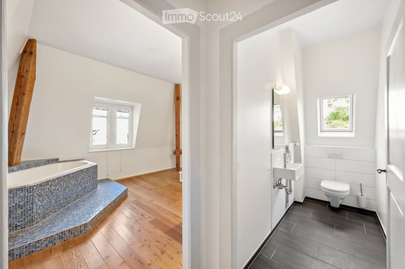 Modern bathroom with a bathtub, sink, and toilet. The space features wooden flooring, white walls, and large windows providing natural light.