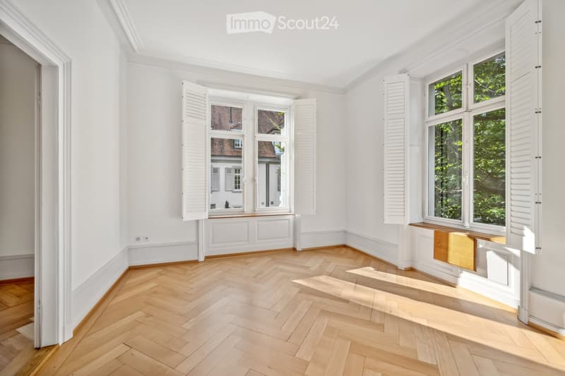 Empty room with white walls, herringbone wooden flooring, large windows with shutters, and a small wooden bench under one window.
