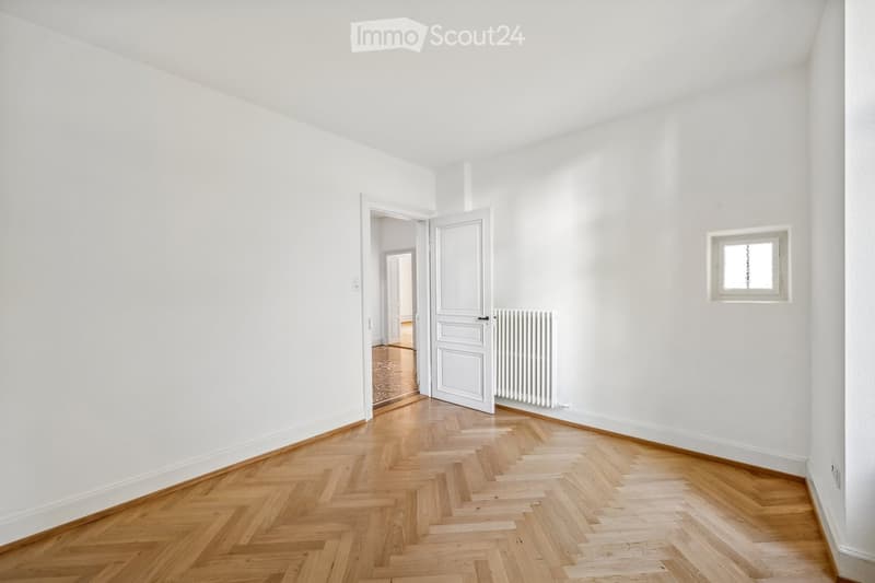 Empty room with white walls, wooden herringbone flooring, a small window, and an open door leading to another room.