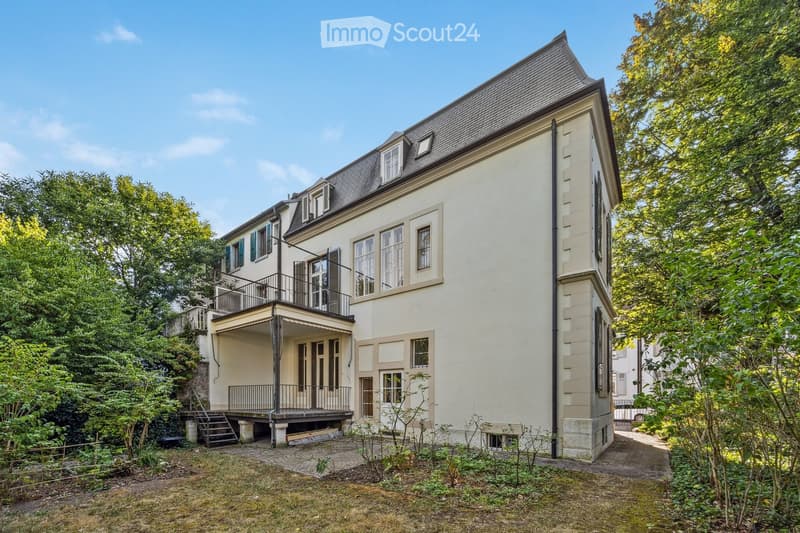 A two-story house with a raised ground floor, featuring a balcony, multiple windows, and a staircase leading to the entrance.