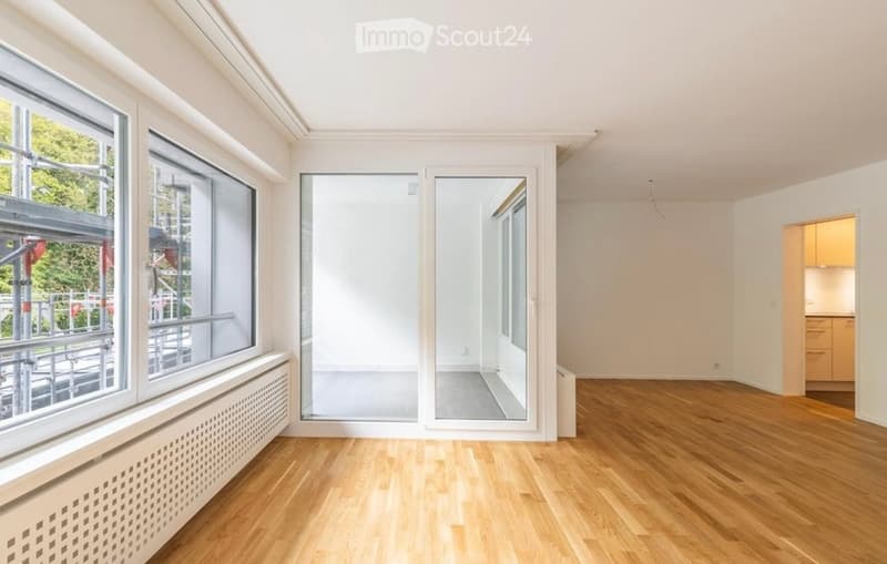 Bright, empty room with wooden flooring, large windows, and a sliding glass door leading to a balcony.