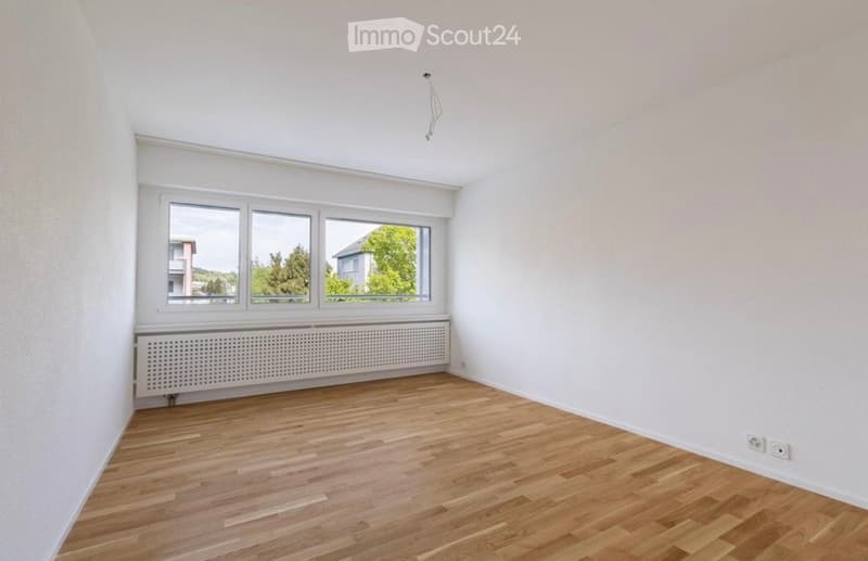 Empty room with wooden flooring, white walls, large windows, and a radiator below the windows.