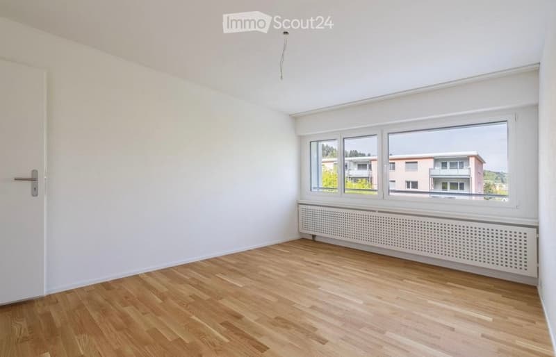 Empty room with wooden flooring, white walls, and large windows offering a view of neighboring buildings.