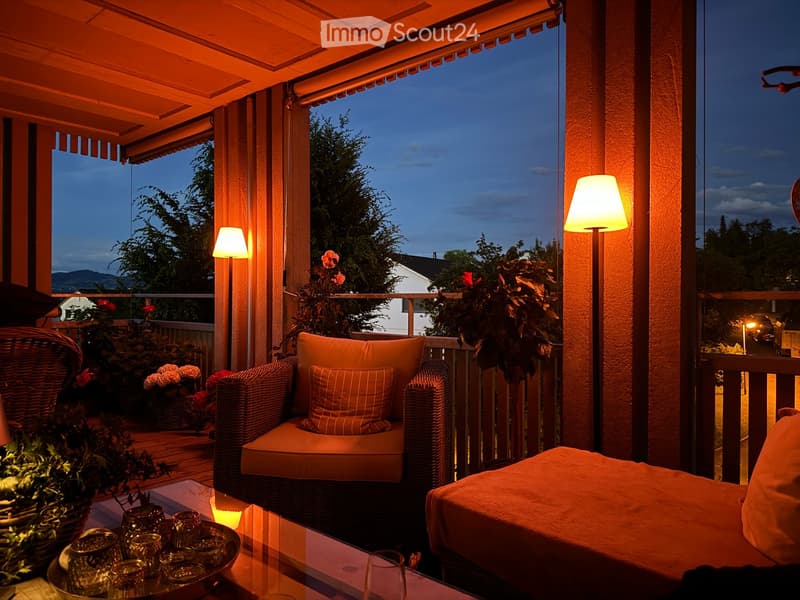 Well-lit balcony with wicker seating, cushions, glass table, and decorative plants, offering a scenic evening view.