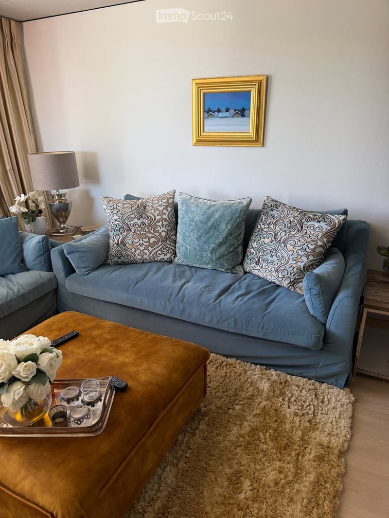 A modern living room with a blue sectional sofa, patterned throw pillows, a mustard-colored coffee table, a shaggy white rug, and a decorative tray with glasses and a remote control. A table lamp and a flower vase are placed on a side table, and a framed
