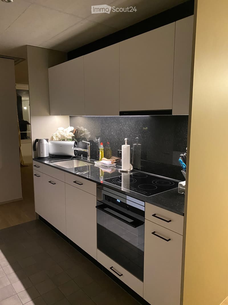 Modern kitchen with light-colored cabinets, black countertops, induction cooktop, sink, and various appliances including a toaster and paper towel holder.