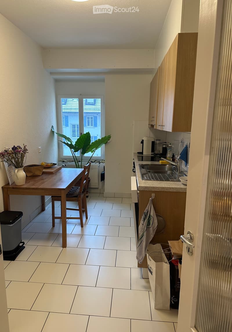 A modern kitchen with white tiled flooring, wooden cabinets, a sink, and a dining area with a wooden table and chairs. A large window offers a view of the outside, and a potted plant adds a touch of greenery.