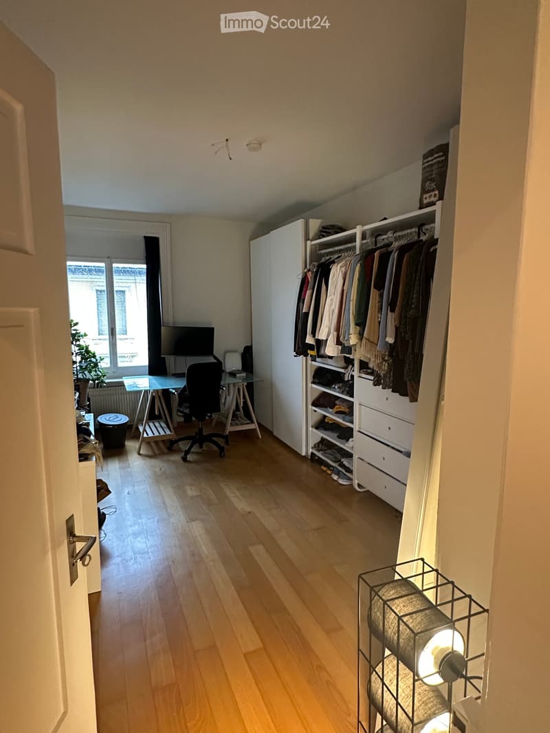 A well-lit room with wooden flooring, a large window, a desk with a chair, a wardrobe with hanging clothes, and a shelf with folded items.