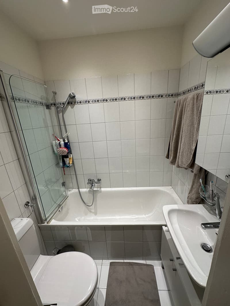 Modern bathroom with a bathtub, toilet, sink, and shower area. Features tiled walls, a towel on the rack, and a toothbrush holder on the sink.