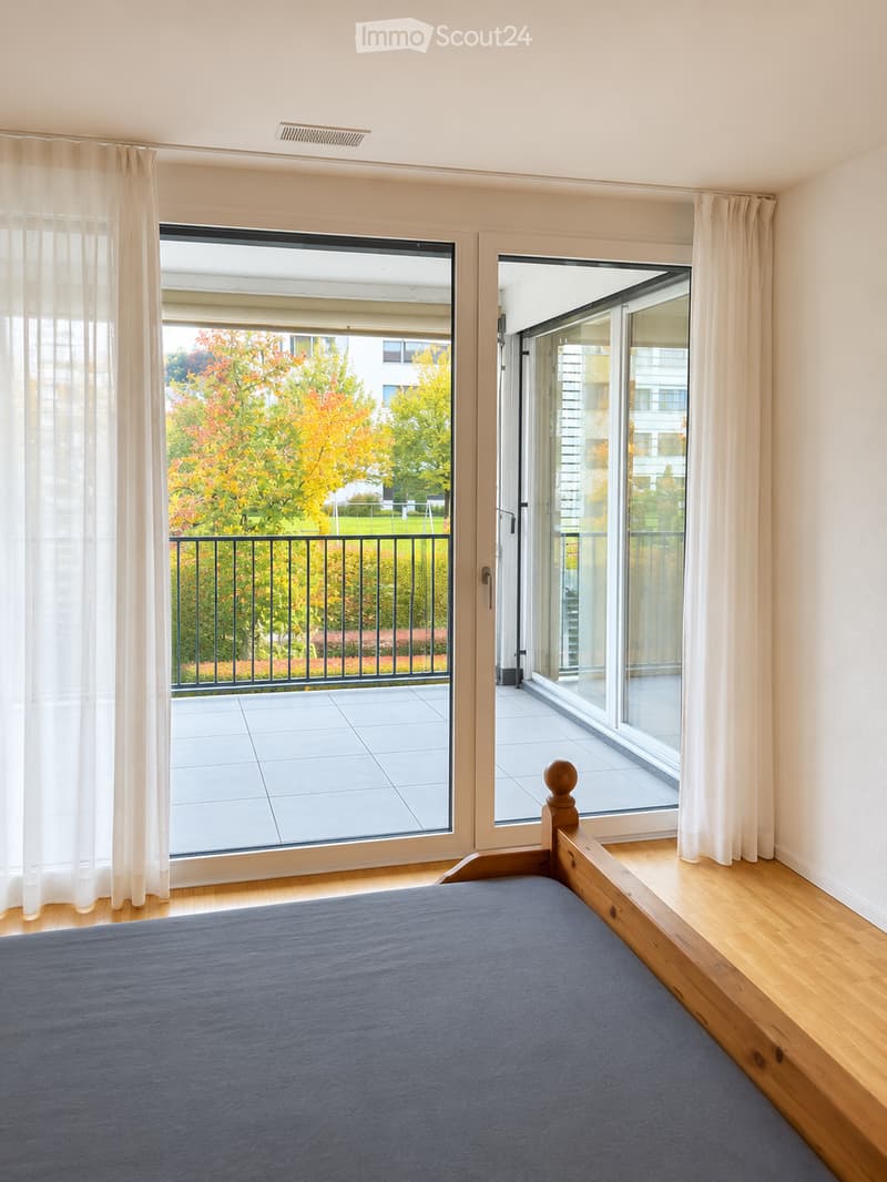 Bright bedroom with wooden flooring, large windows leading to a balcony, and a neatly made bed.