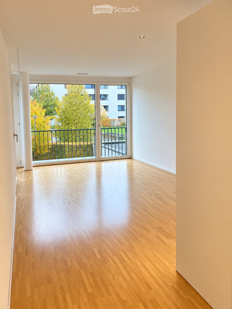 Empty room with light wooden flooring, white walls, and large sliding glass doors leading to a balcony with a metal railing and view of greenery.