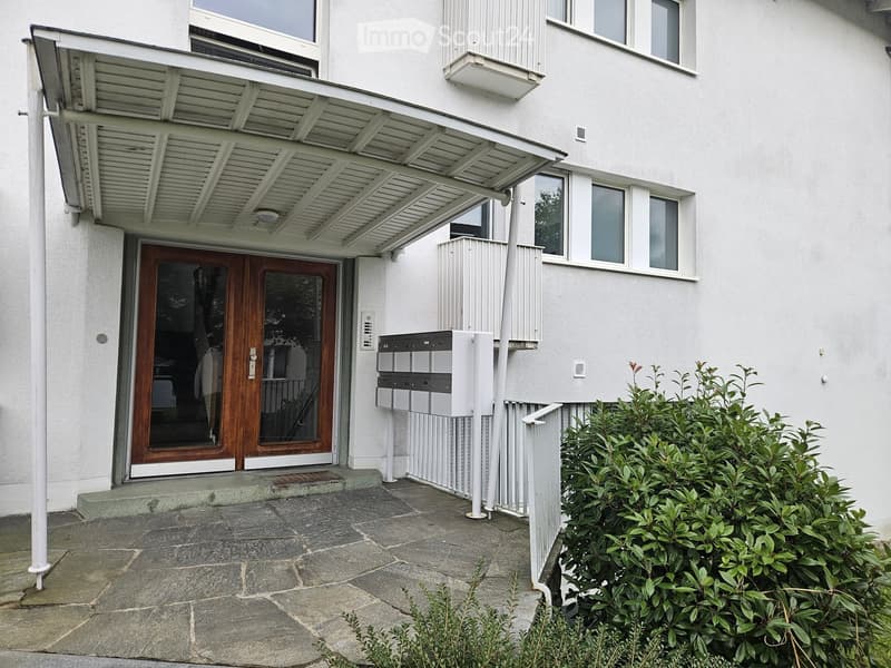 Modern building entrance with a covered porch, wooden double doors, mailboxes, and small garden area.