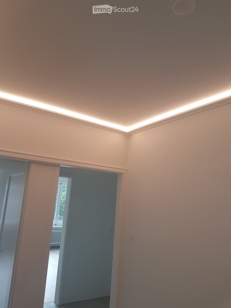 Empty hallway with beige walls, white ceiling lighting, and a partially open door leading to another room.