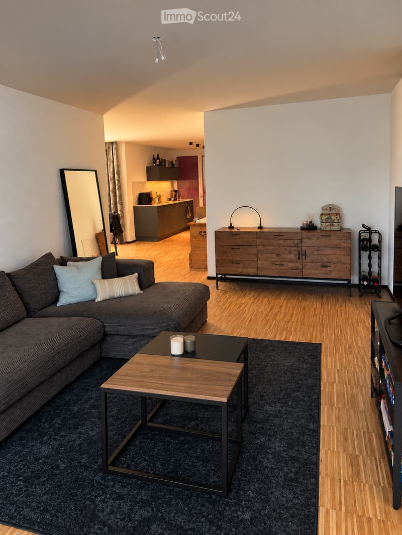 Modern living room with a sectional sofa, coffee table, and a large mirror. The room features a wooden dresser, a wine rack, and a television. An open kitchen area is visible in the background with cabinets, a counter, and various appliances.
