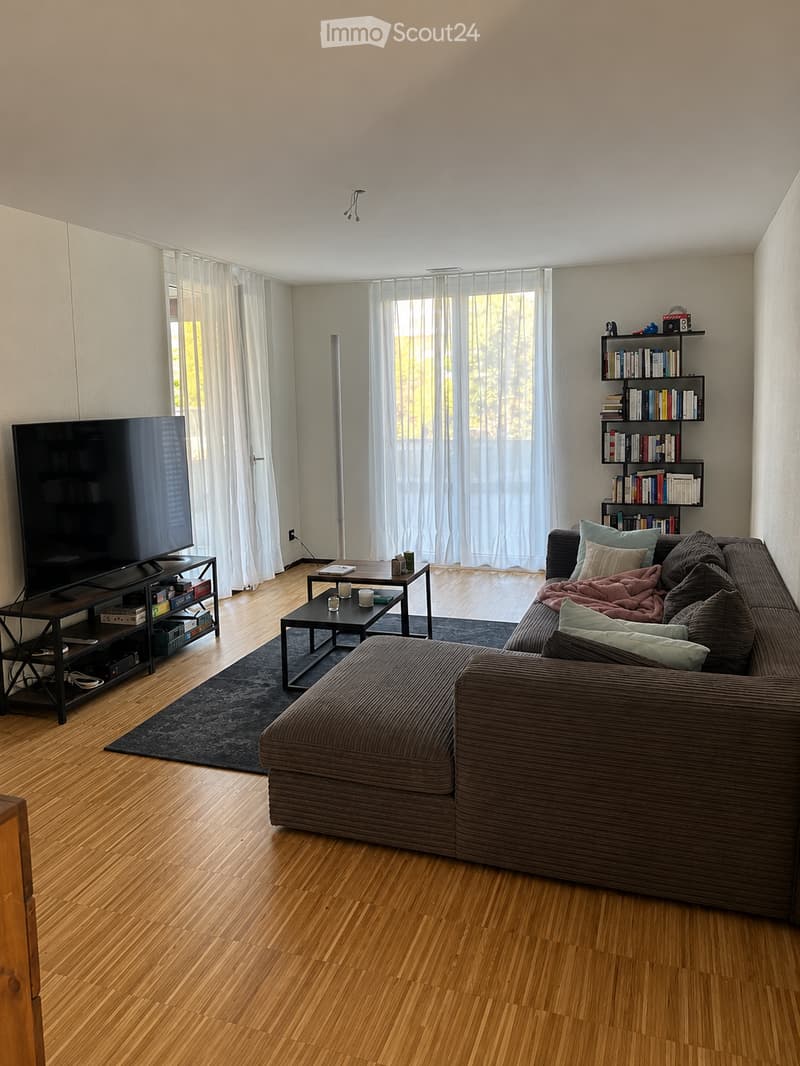 Modern living room with wooden flooring, large windows with white curtains, a sectional sofa, coffee table, TV stand, bookshelf, and a small table with candles.