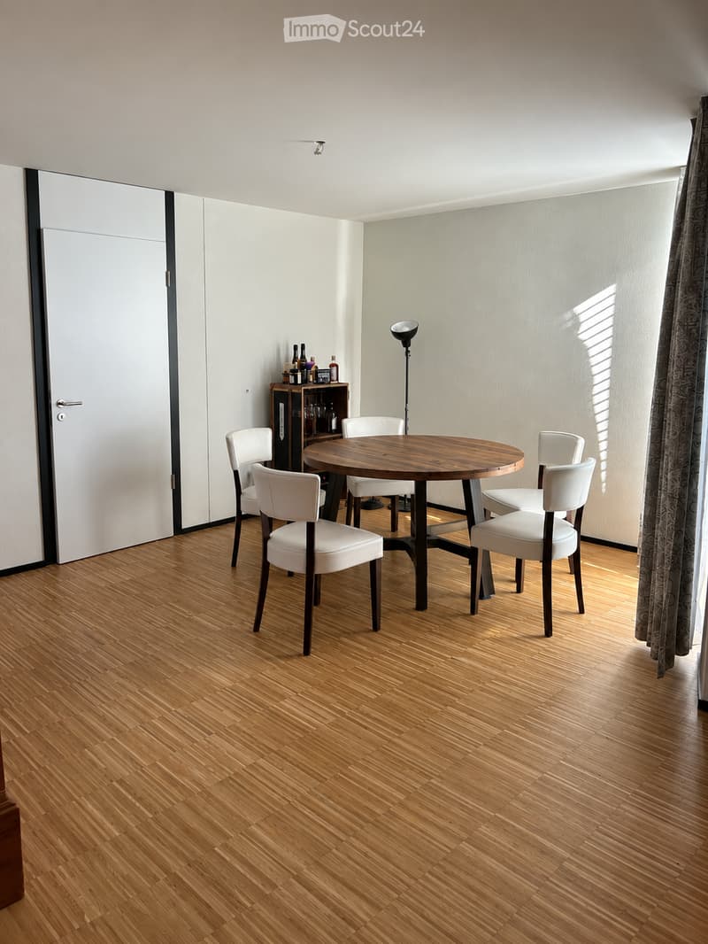 A modern dining area with a round wooden table surrounded by white chairs, a small wooden shelf with bottles, and a floor lamp near a large window with curtains.