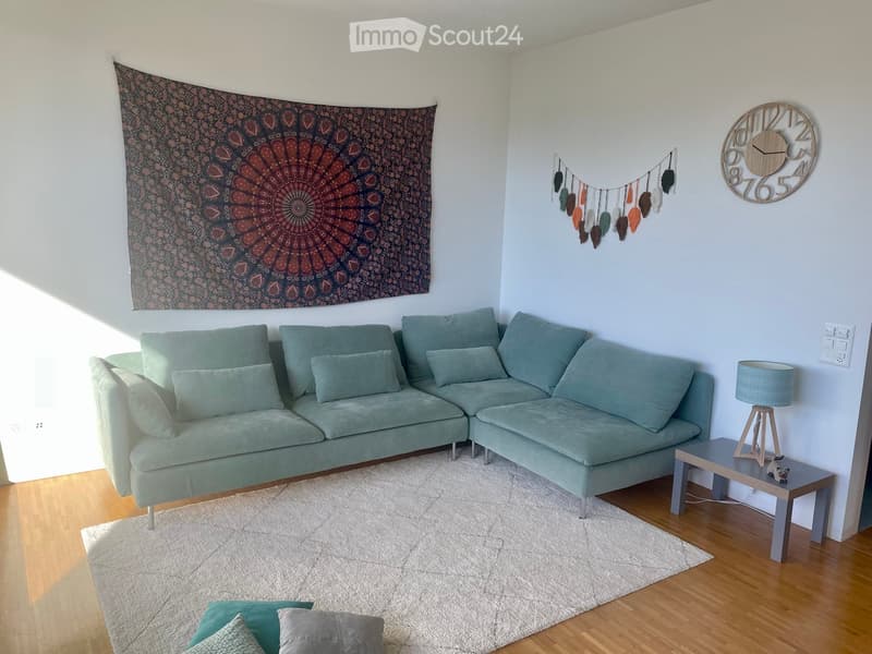 Modern living room with a light green sectional sofa, wooden coffee table, table lamp, wall tapestry, and decorative hanging.