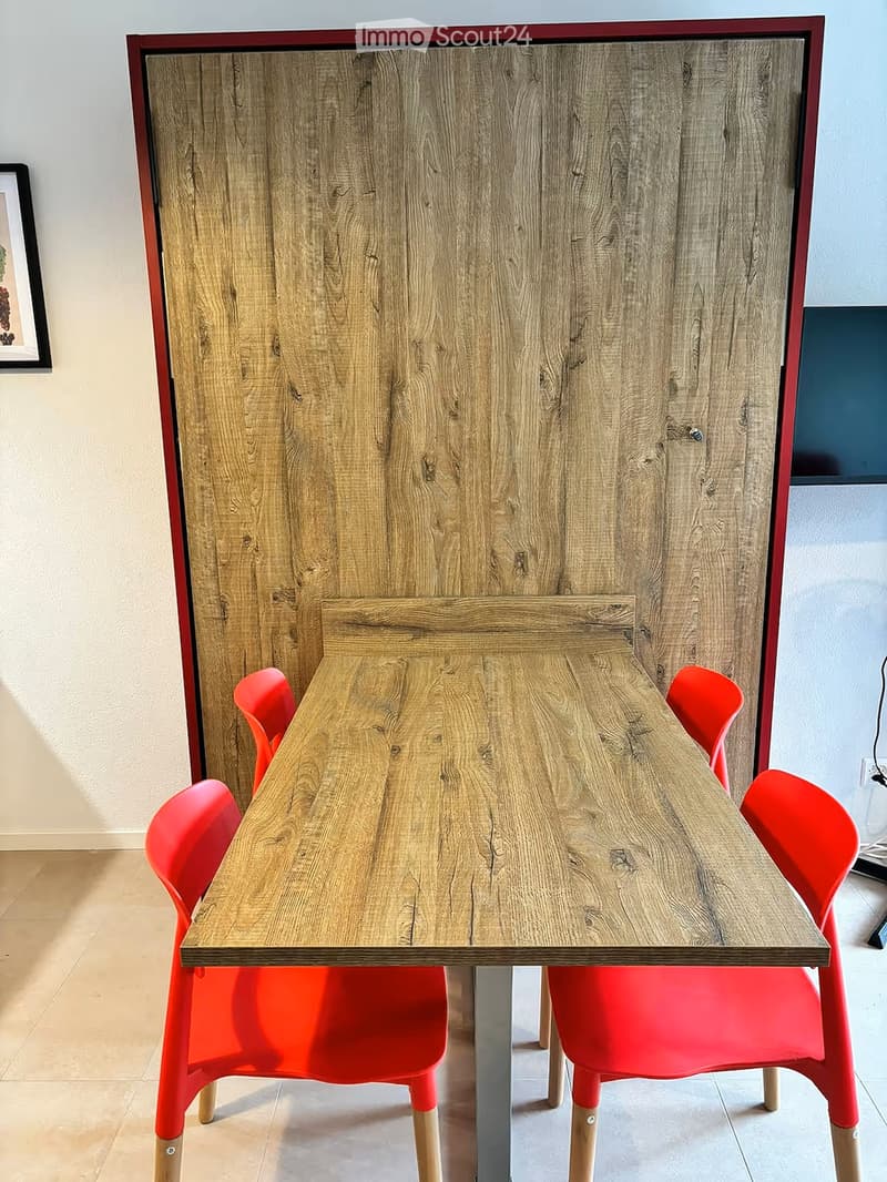 A modern dining area with a wooden table and four red chairs, set against a light-colored wall with a framed picture and a large wooden cabinet.