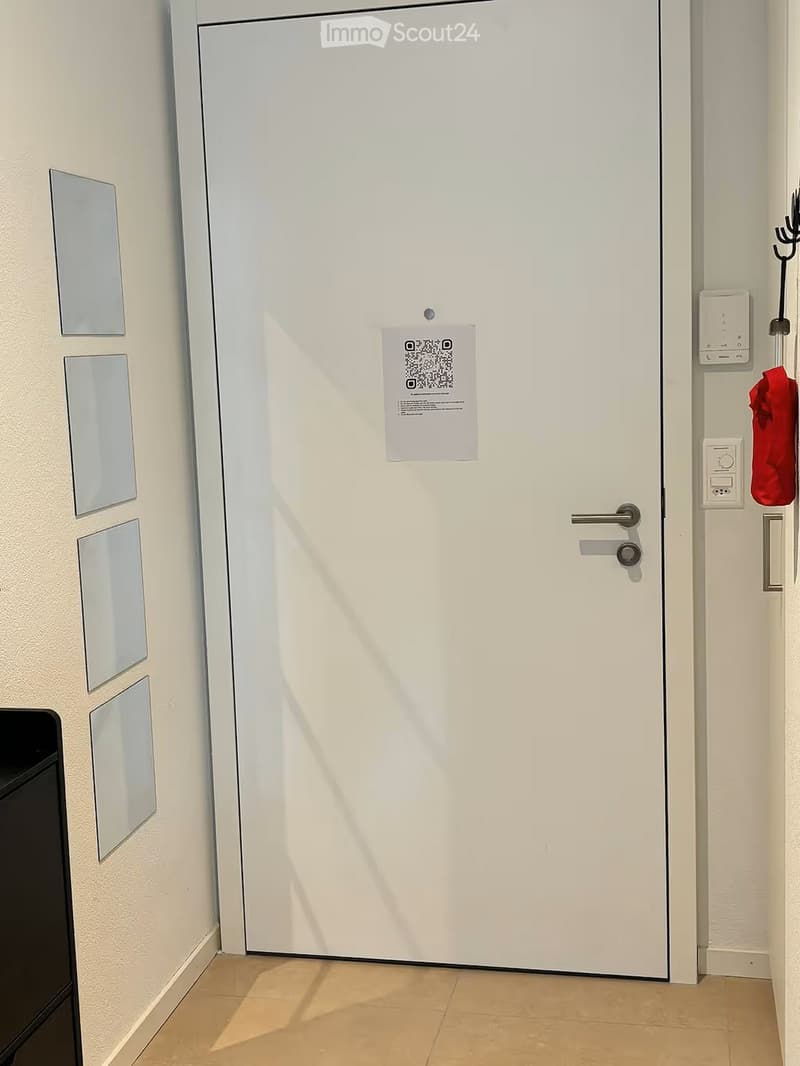 A hallway with a white door featuring a QR code sticker, a coat hook with a red coat, and a light switch panel on the wall.