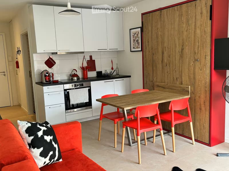 Modern open-plan living and dining area with a red sofa, a wooden dining table with red chairs, a kitchen area with white cabinets, an oven, and a fridge.