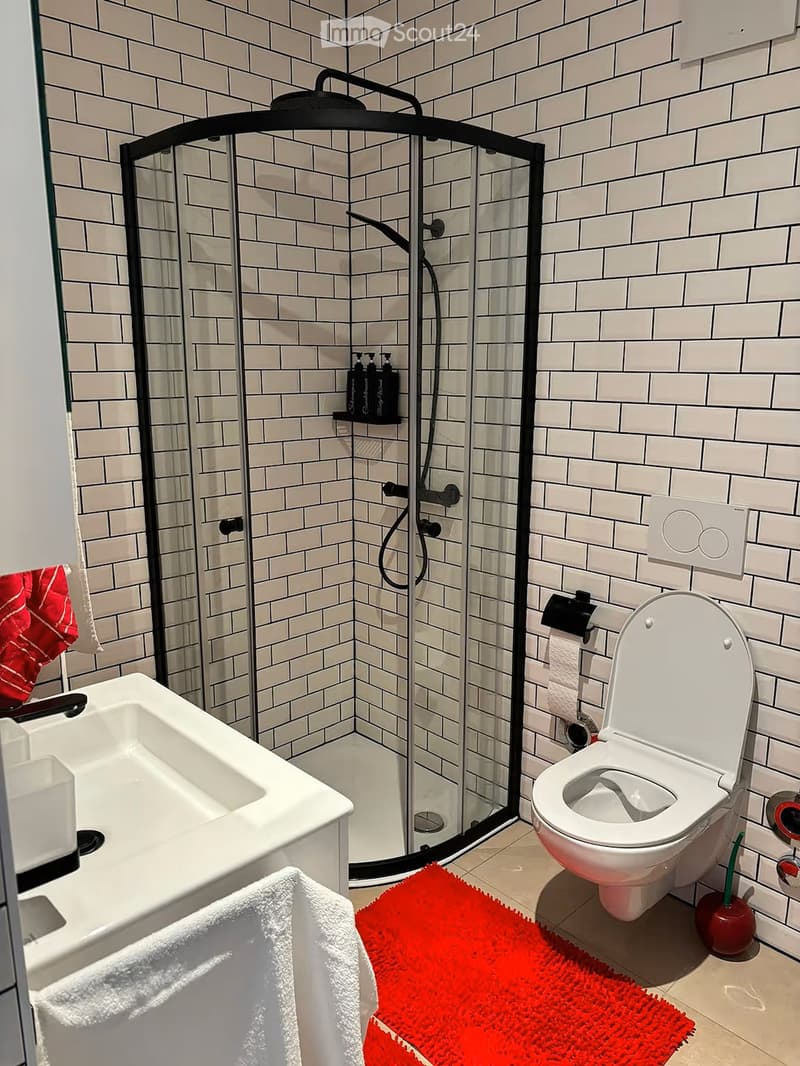 Modern bathroom with white tiles, a glass-enclosed shower, a toilet, a sink, and a red towel on the floor.