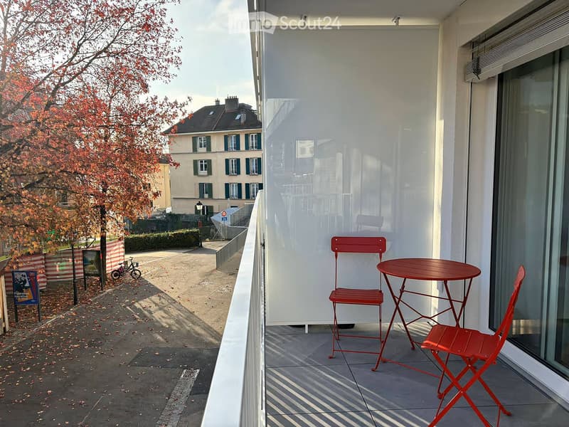 Balcony with a red table and two red chairs, overlooking a street with trees and buildings.