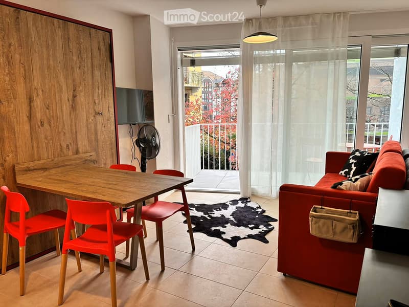 A modern living room with a red sofa, wooden dining table, red chairs, a fan, and a view of a balcony with outdoor furniture.
