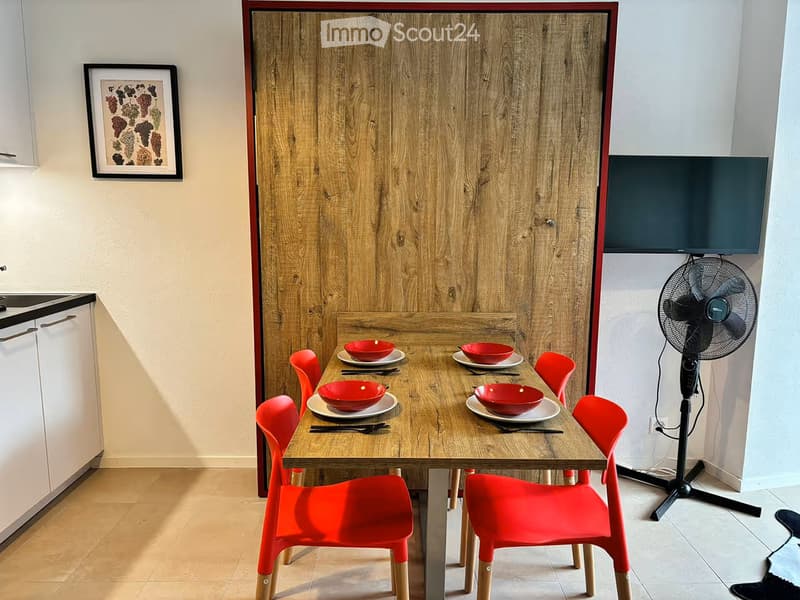 A modern dining area with a wooden table, red chairs, and a fan. The table is set with red bowls and plates, and there is a screen mounted on the wall.