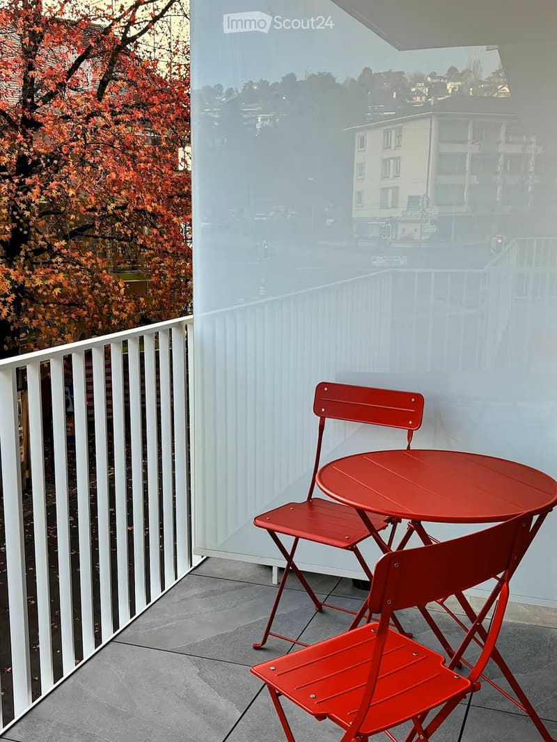 Balcony with a red folding table and two matching chairs, white railing, and a view of the street and surrounding buildings.