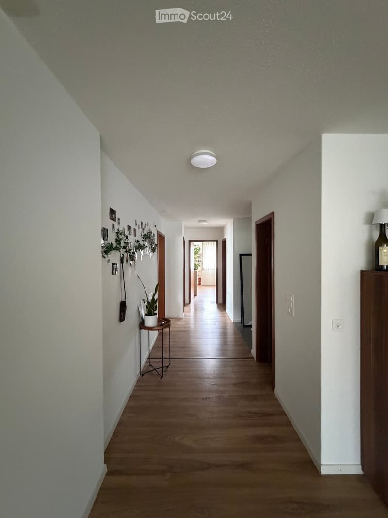 A long, well-lit hallway with wooden flooring, white walls, and several doors leading to different rooms. A small side table with a potted plant is visible on the left, and a lamp is placed on a wooden shelf on the right.