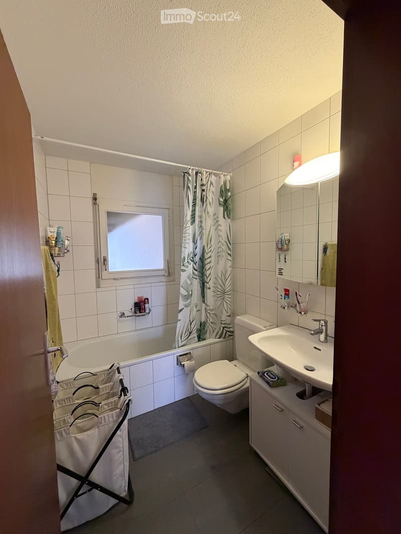Modern bathroom with white tiles, bathtub, toilet, sink, mirror, and a shower curtain with a leaf pattern. Includes a towel rack, toothbrush holder, and a small cabinet under the sink.