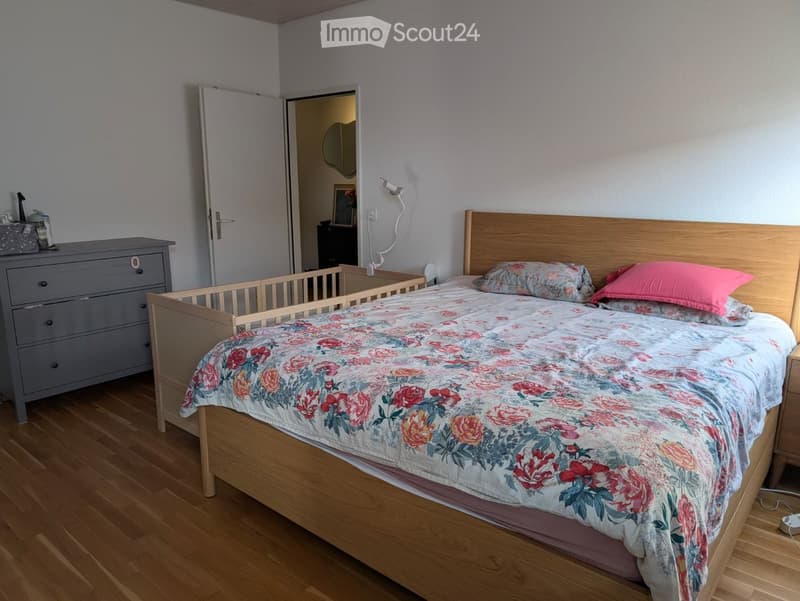 Cozy bedroom with a wooden bed, floral duvet, pink pillows, nightstand, dresser, and a mirror in the adjacent room.