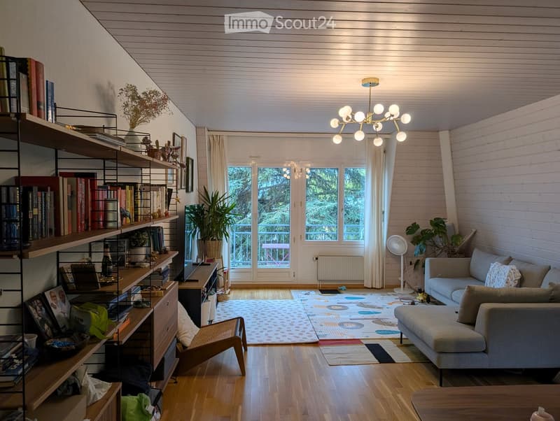 Bright living room with wooden floors, white paneled ceiling, large windows, and a view of greenery. Features a grey sofa, coffee table, bookshelf, and a small fan.