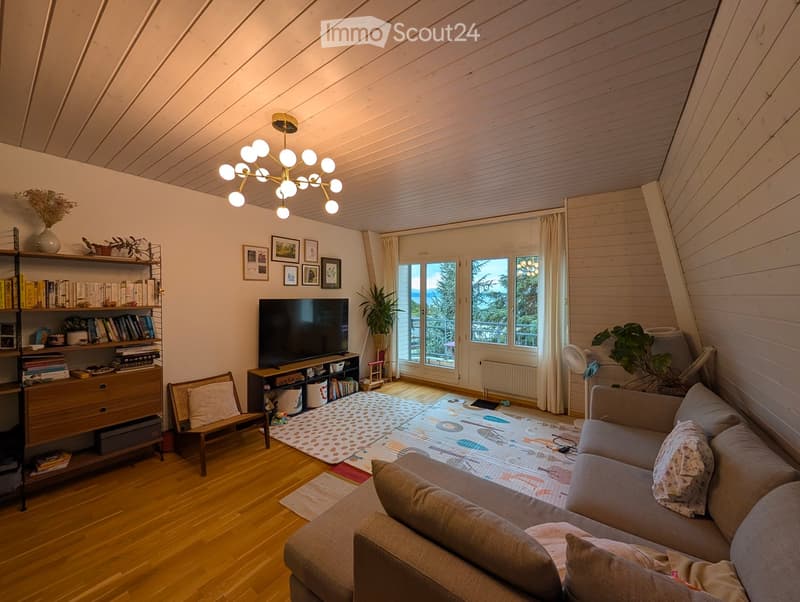 Modern living room with wooden floors, a grey sectional sofa, coffee table, bookshelf, TV, and large windows with a view.